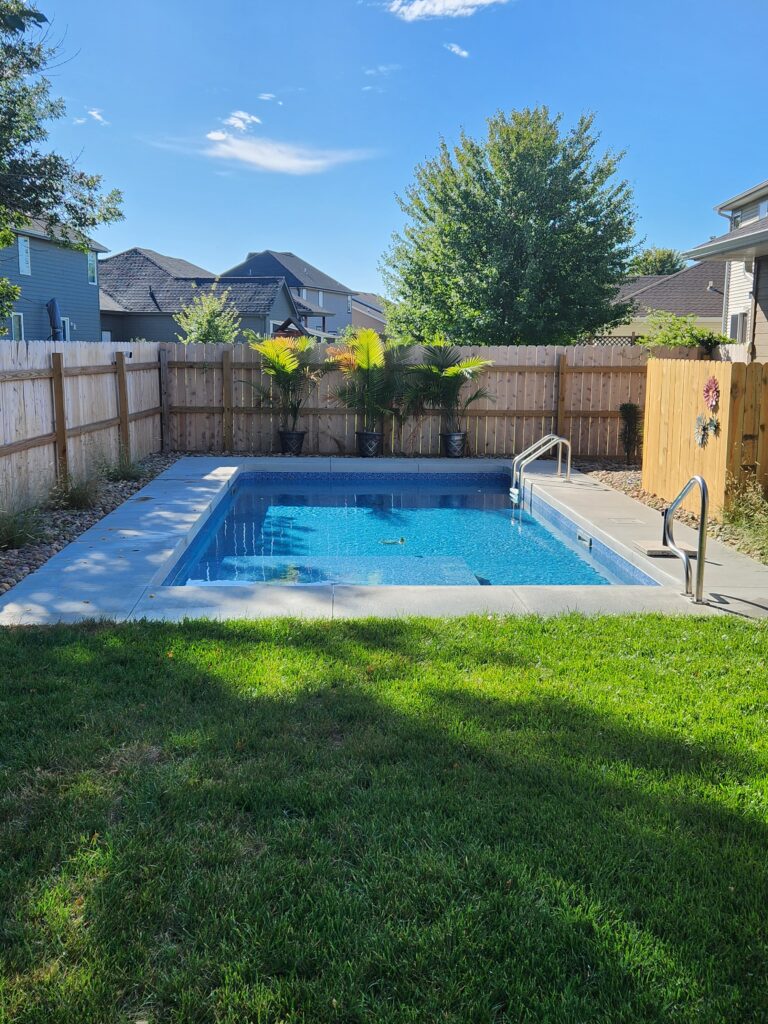 A small rectangular New Pool with clear blue water is surrounded by a wooden fence and greenery, with steps leading into the pool and a patch of grass in the foreground—another beautiful creation by Dreamscape Pools.