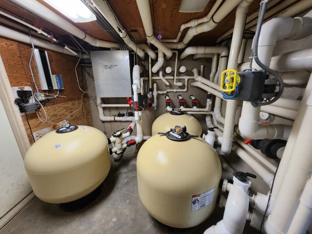 A utility room for Dreamscape Pools, featuring large round beige filtration tanks, intricate white PVC pipes weaving through the space, and electrical control boxes on wooden walls. The network of pipes and valves adds to the complexity.