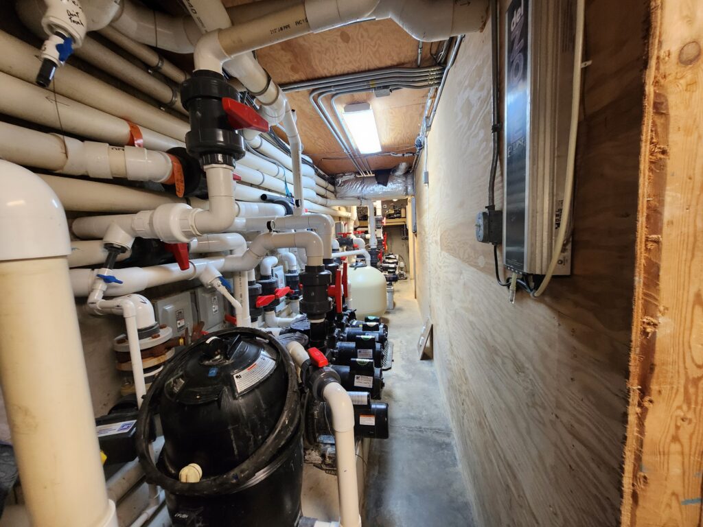 Narrow utility room lined with numerous white pipes, valves, electrical wiring, pumps, and Dreamscape Pools filtration equipment attached to the walls and ceiling. The space has a wooden wall on one side and appears organized but busy.