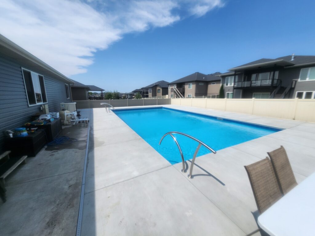 A rectangular outdoor swimming pool by Dreamscape Pools features clear water, a concrete deck, and patio furniture, set among residential houses under a partly cloudy blue sky, perfect for your new pool retreat.