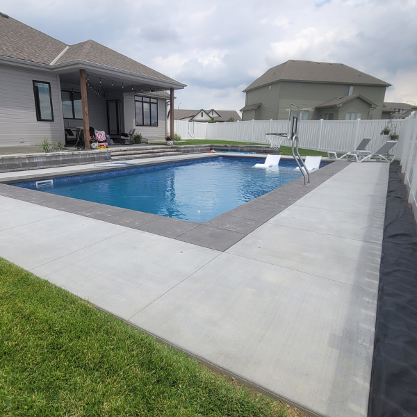 A backyard swimming pool with gray stone edging, white lounge chairs in the water, and a covered patio area attached to a house; fenced-in yard with grass and cloudy sky overhead.
